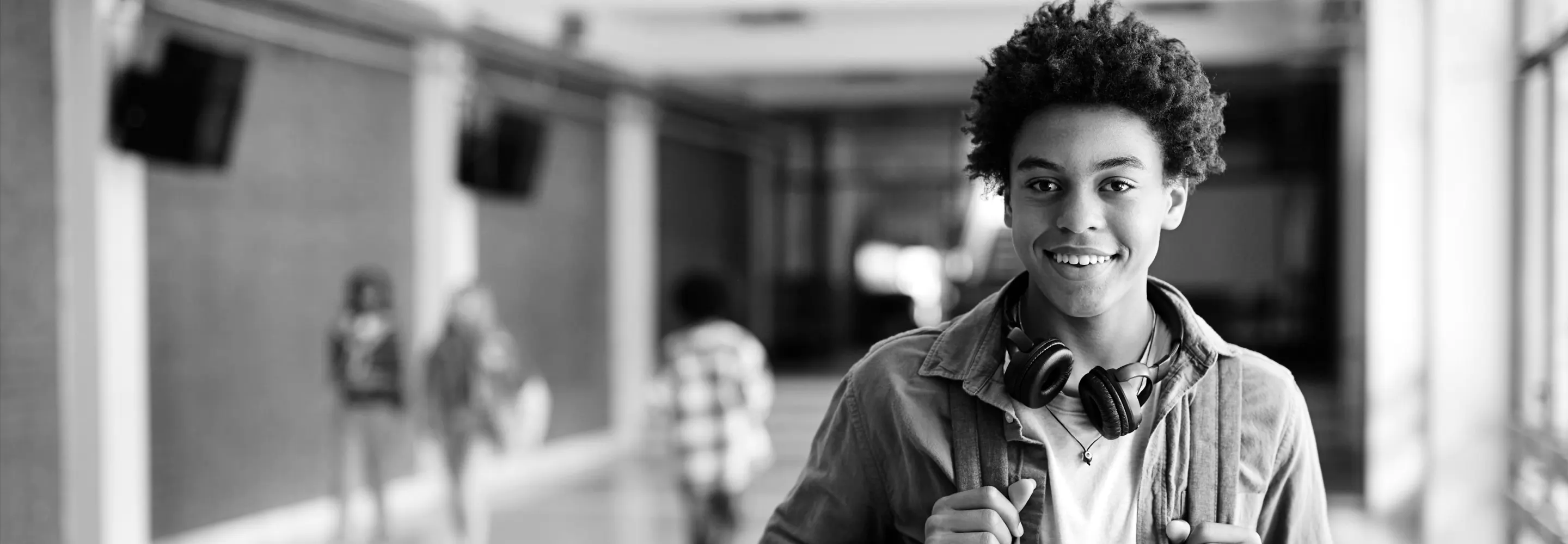 A smiling high school student with headphones around his neck stands in a school hallway
